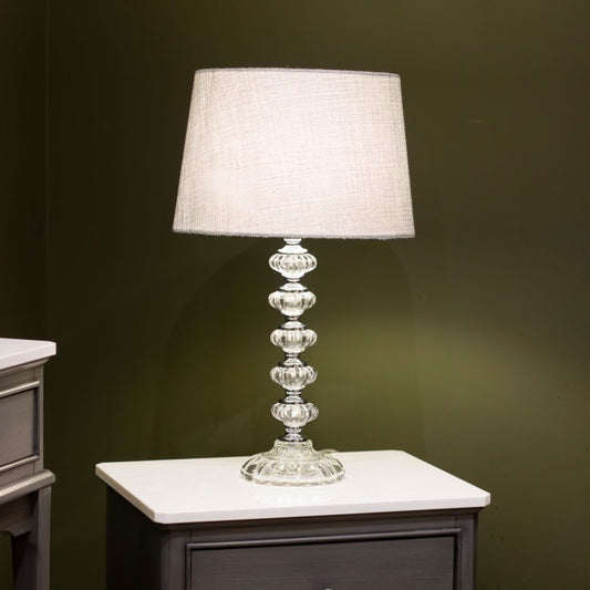 Decorative table lamp with a clear glass base and grey lampshade on a dark surface.