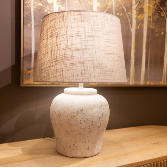 Table lamp with textured shade on a wooden surface, with a painting of trees in the background.