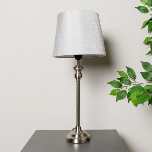 Table lamp with a grey shade on a greysurface, with green leaves in the background.